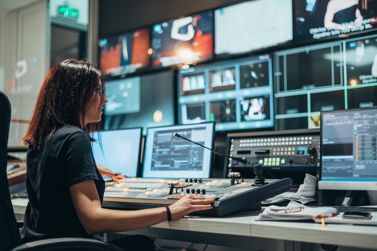 Gallery operator at a broadcast control desk in front of a wall of monitors